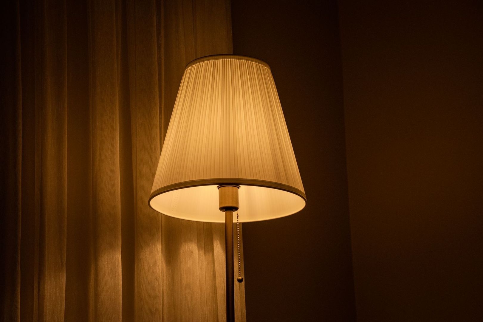 Varmt lysande golvlampa med veckad skärm framför mörka gardiner